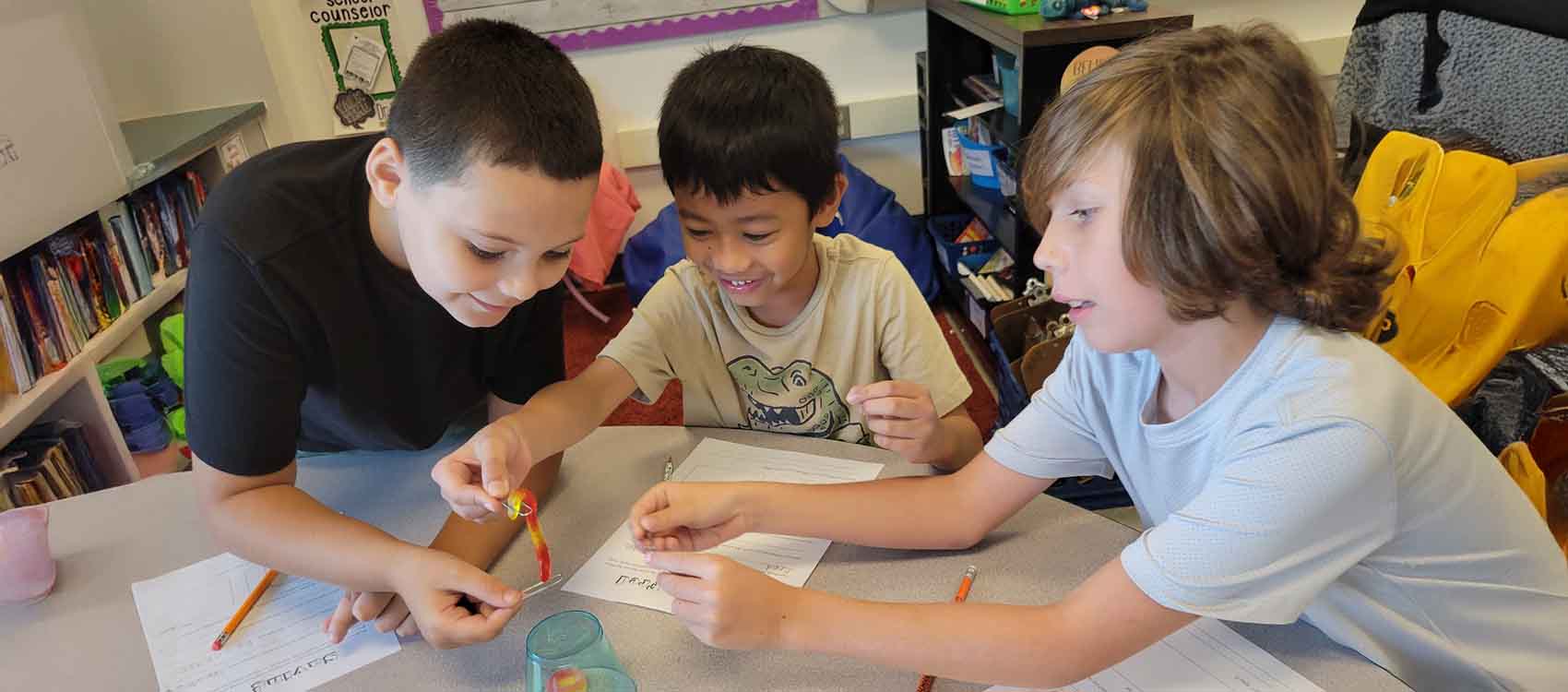 Three elementary school students working together on a hands-on science experiment in a classroom. The student on the left in a black shirt is holding what appears to be a small strip or test material, while the middle student in a beige shirt with a graphic design watches with interest. The student on the right in a light blue shirt is leaning in to observe. They're seated at a table with papers, pencils, and what looks like a small container or beaker. Behind them are classroom elements including bookshelves, storage areas, and educational posters on the walls, creating a typical elementary classroom learning environment.