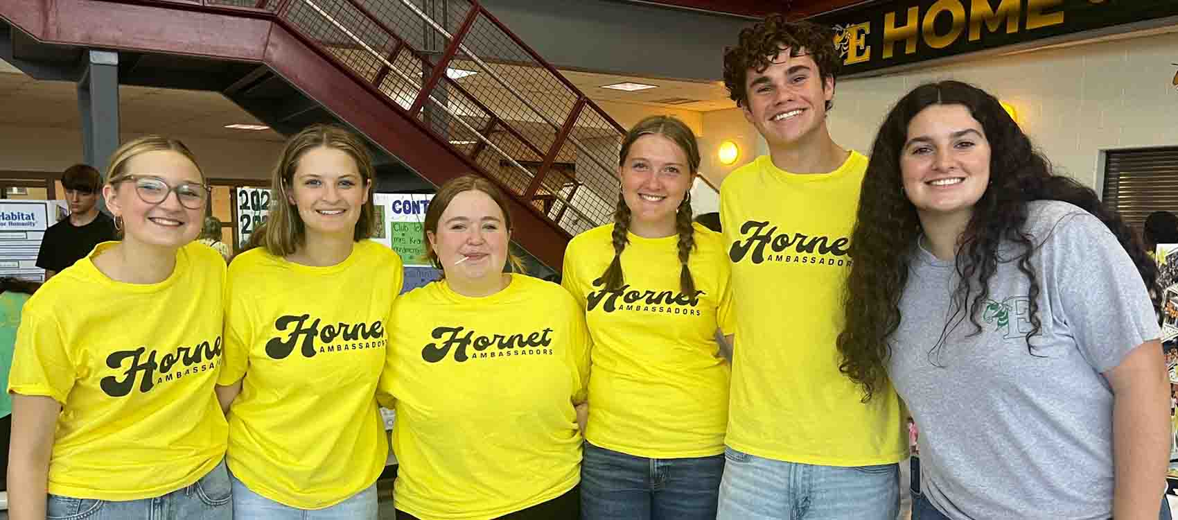 A group photo of six smiling high school students standing together in a school hallway. Five students are wearing matching bright yellow t-shirts that read "Hornet Ambassadors" in black script lettering, while one student on the right wears a gray sweatshirt. The students appear to be part of a student leadership or ambassador program. Behind them is a school staircase with metal railings and partial signage visible on the walls, including what appears to be "HOME" text in the background.