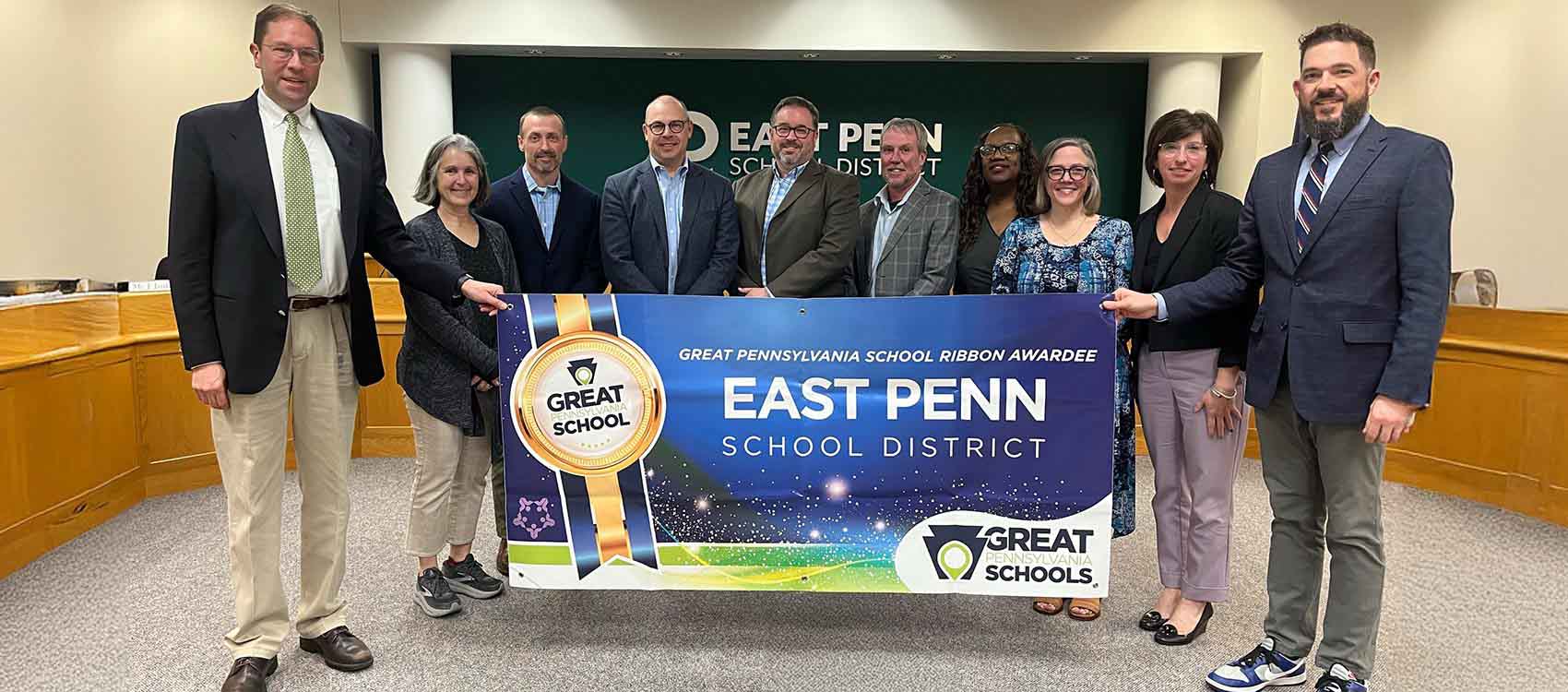 A group of individuals is posing for a photo in front of a banner that reads "Great Pennsylvania School Ribbon Award Awardee East Penn School District." The banner features a blue and gold design with a ribbon and stars. The setting appears to be an indoor office or meeting room