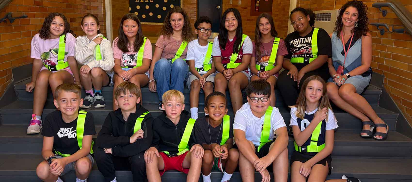 A group of children and an adult are sitting on steps, wearing bright green safety vests. They are smiling and posing for a photo. The background includes a brick wall and a staircase.