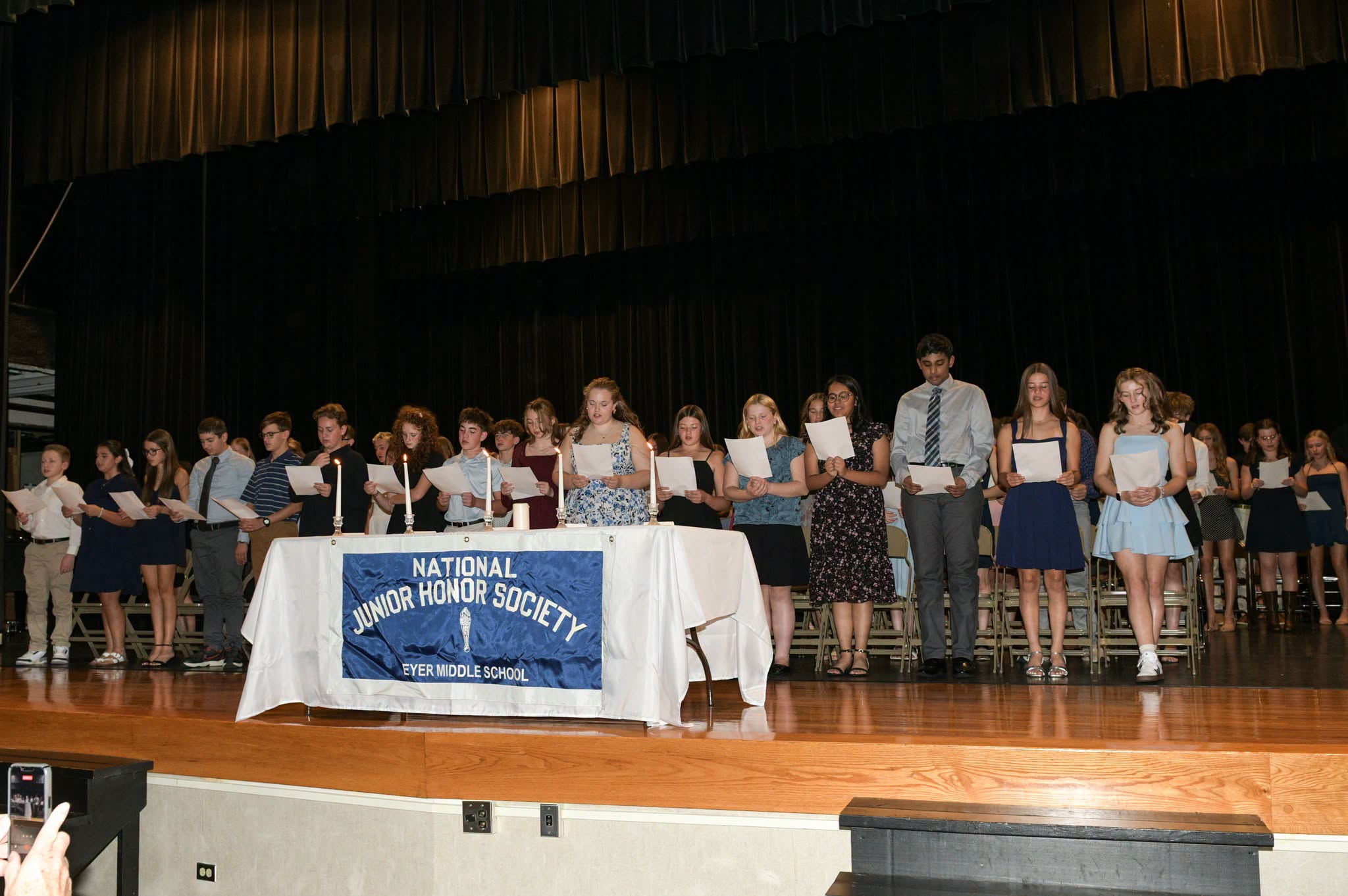A wide shot of approximately 18 middle school students standing side-by-side on a polished wooden stage in front of a black curtain. They are dressed in semi-formal clothing—dresses and button-down shirts—and most are holding white sheets of paper. In front of them is a table covered with a white cloth featuring a blue banner that reads "NATIONAL JUNIOR HONOR SOCIETY" and "EYER MIDDLE SCHOOL."