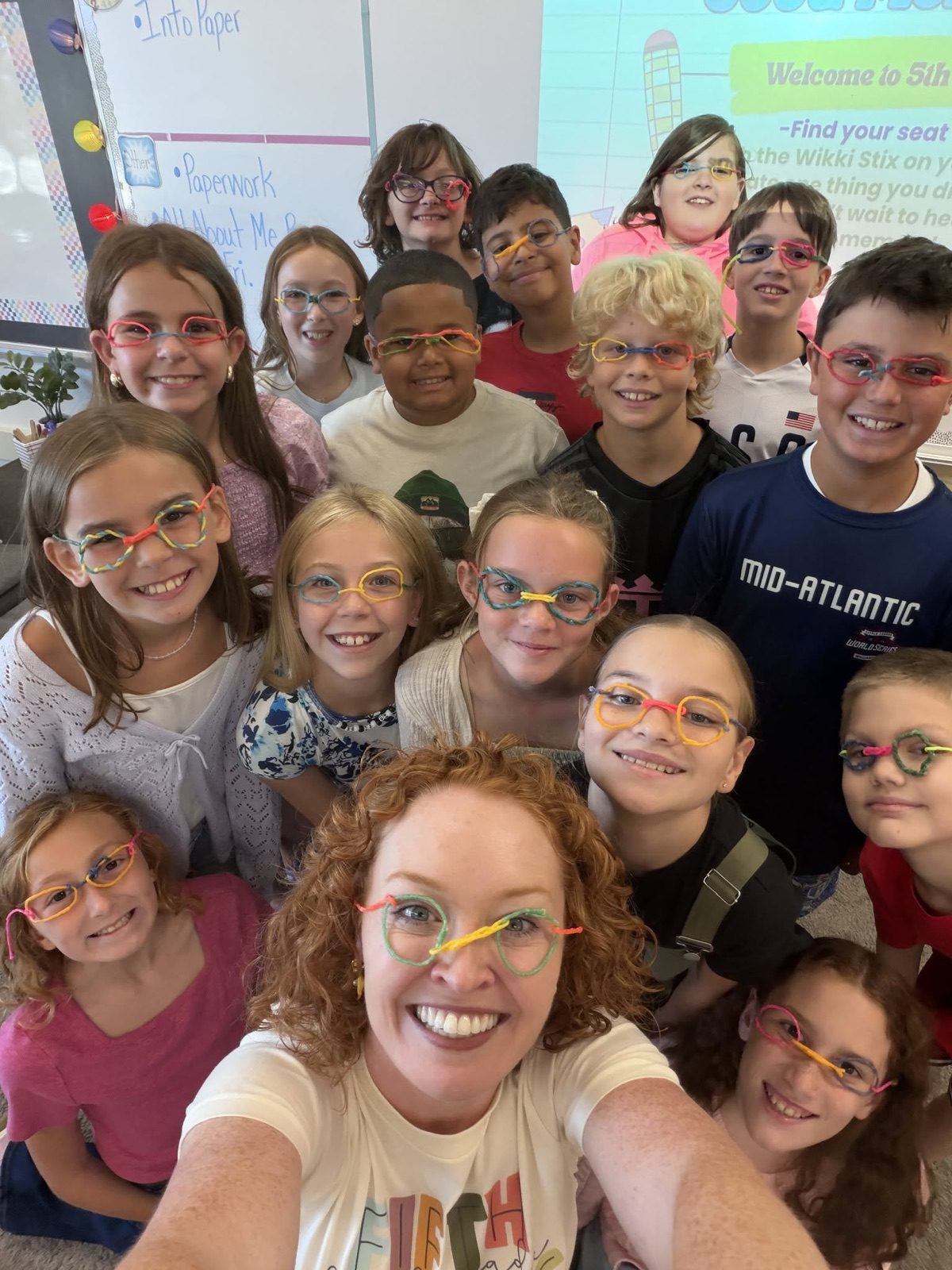 A teacher takes a selfie with her smiling class, everyone wearing colorful twist-straw glasses