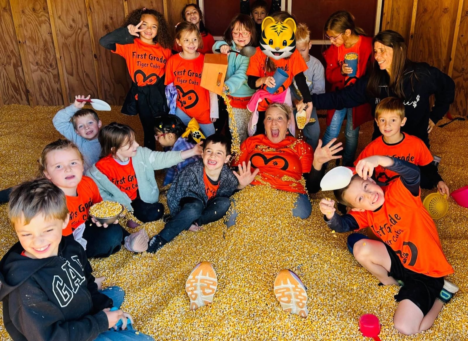 A first-grade class in matching orange shirts laughs and plays in a corn pit with their teachers and a tiger mascot