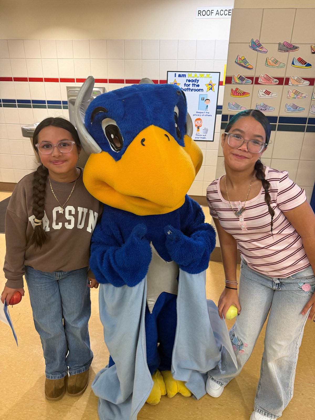 Two students smile beside their school’s blue bird mascot in a school hallway