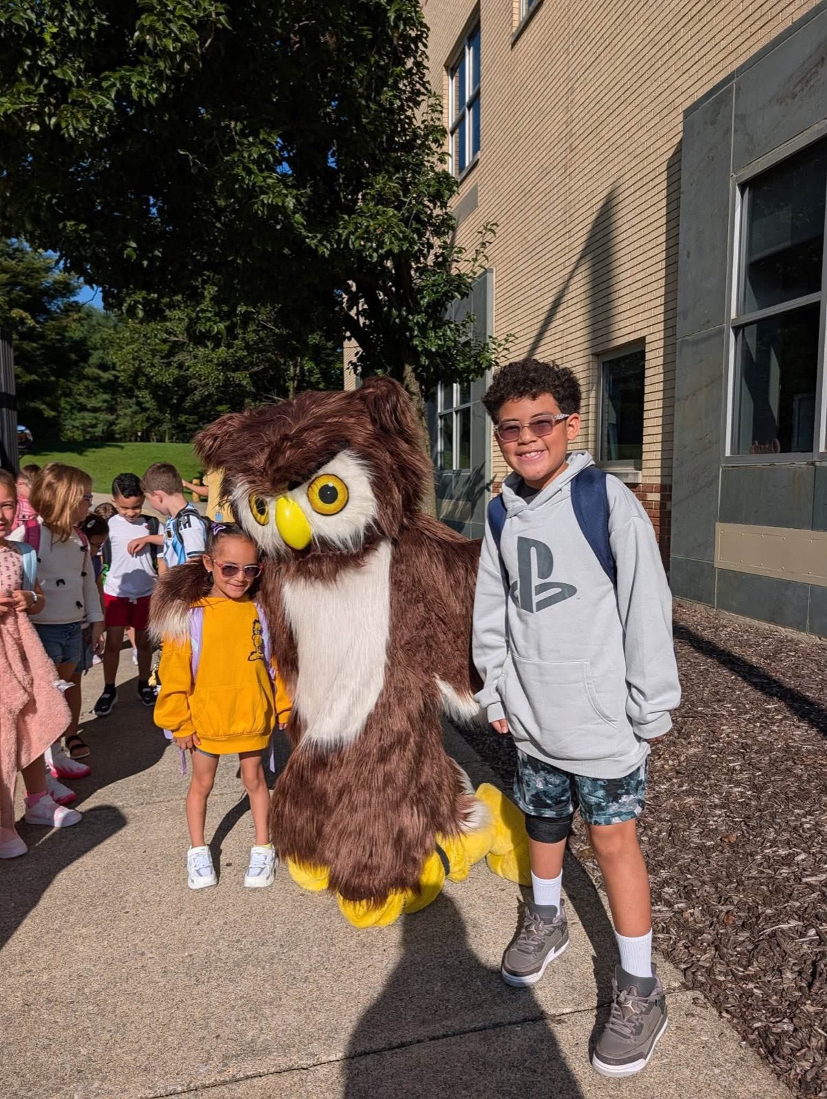 Two smiling elementary students stand beside their school’s owl mascot outside the building