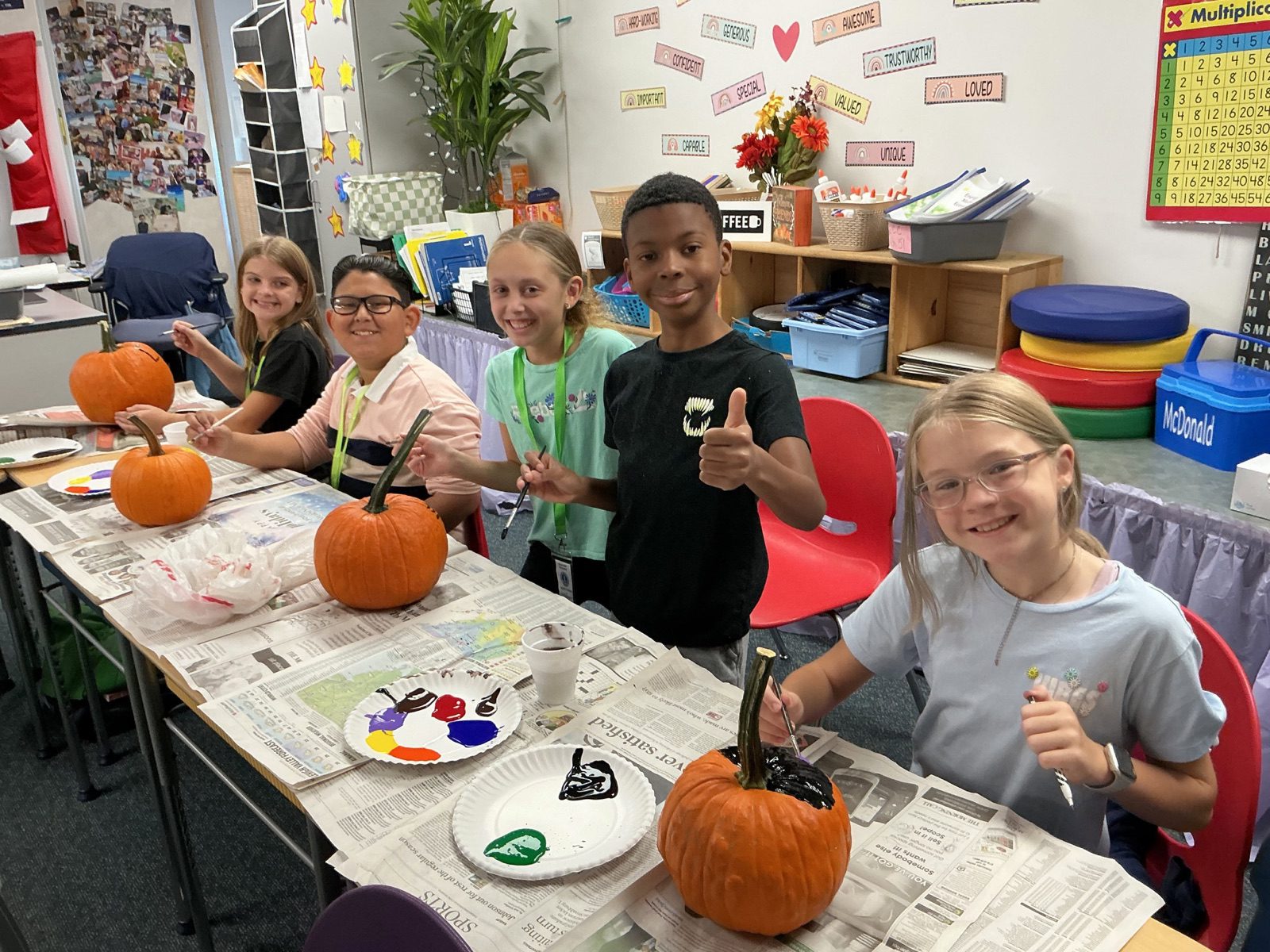 Elementary students smile and give a thumbs-up while painting pumpkins in their classroom